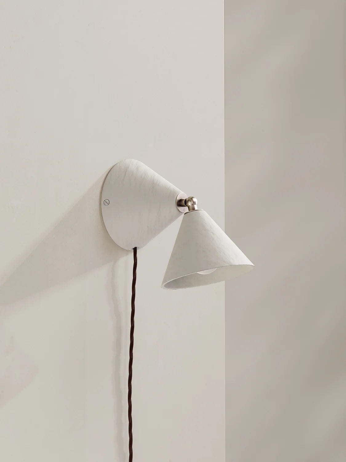 A modern white wall-mounted cone-shaped sconce with a dark brown cord, shown mounted on a minimalist off-white wall.