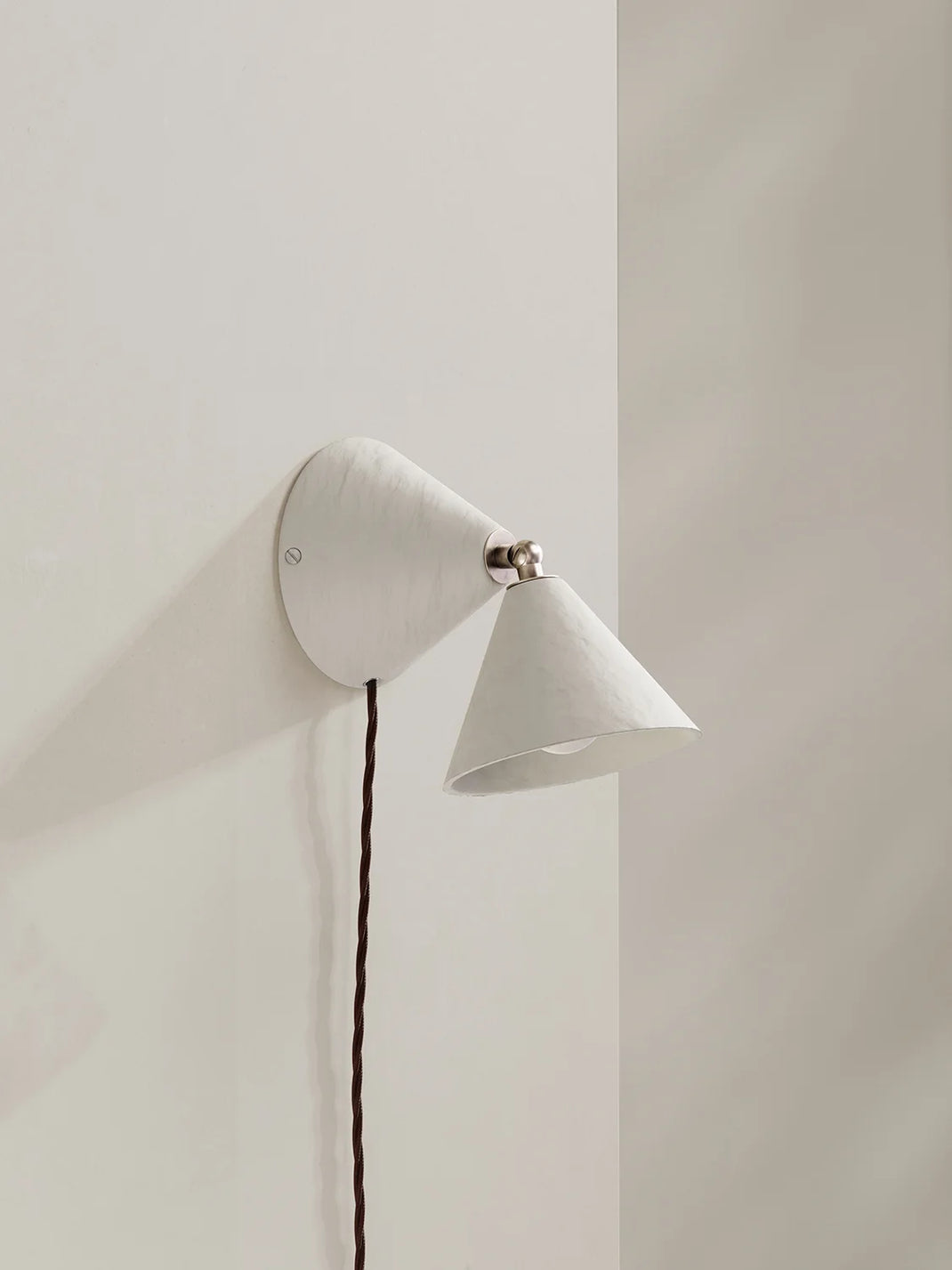 A modern white wall-mounted cone-shaped sconce with a dark brown cord, shown mounted on a minimalist off-white wall.