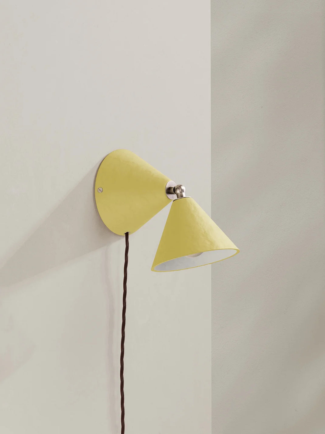 A modern wall-mounted cone-shaped light fixture in a soft muted yellow, shown mounted on a neutral off-white wall with its dark brown cord hanging down.