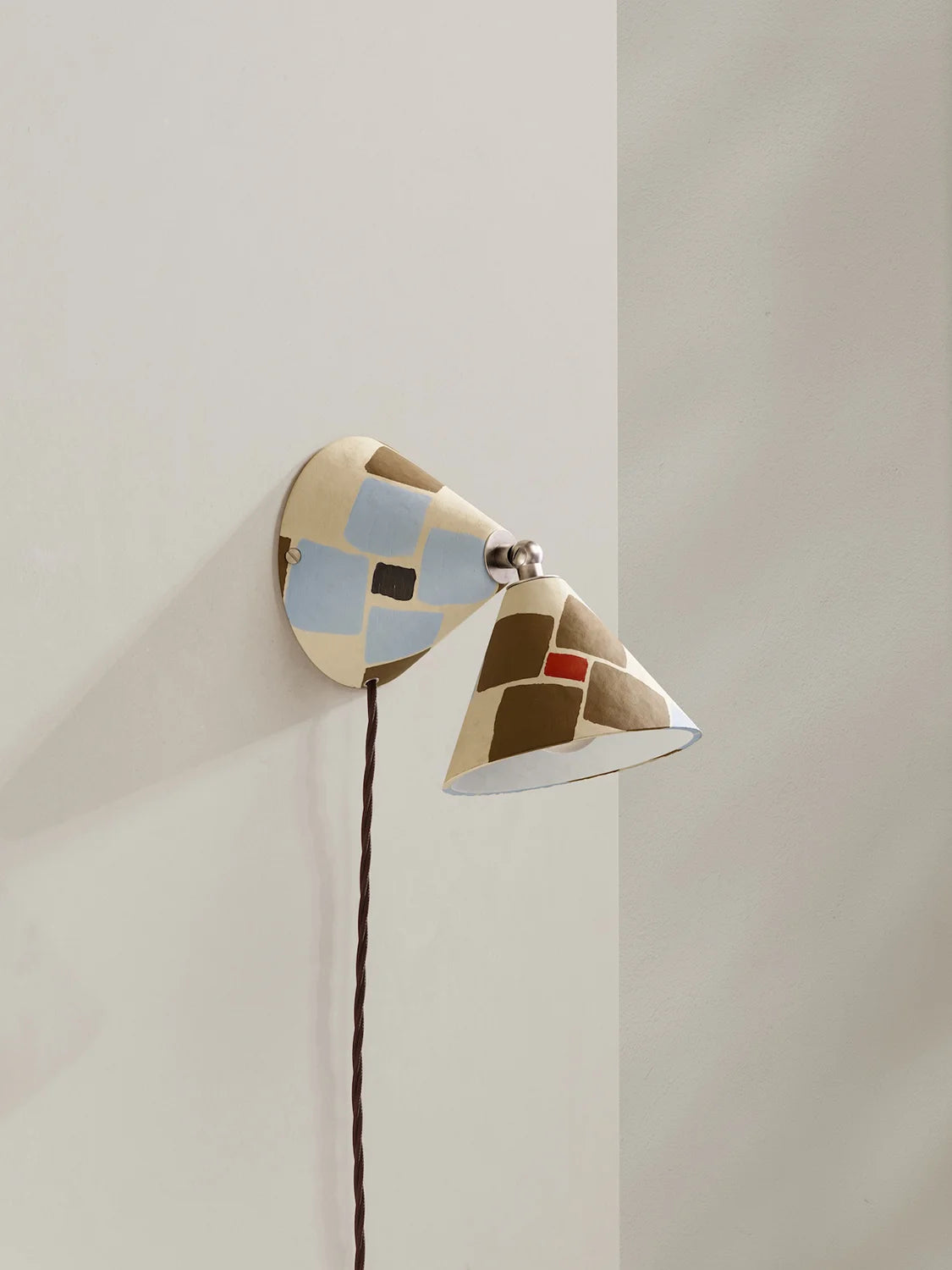 A modern wall-mounted sconce with a dual-cone design, featuring a cream-colored body with geometric patches of light blue, brown, and a small red accent, mounted on a beige wall.