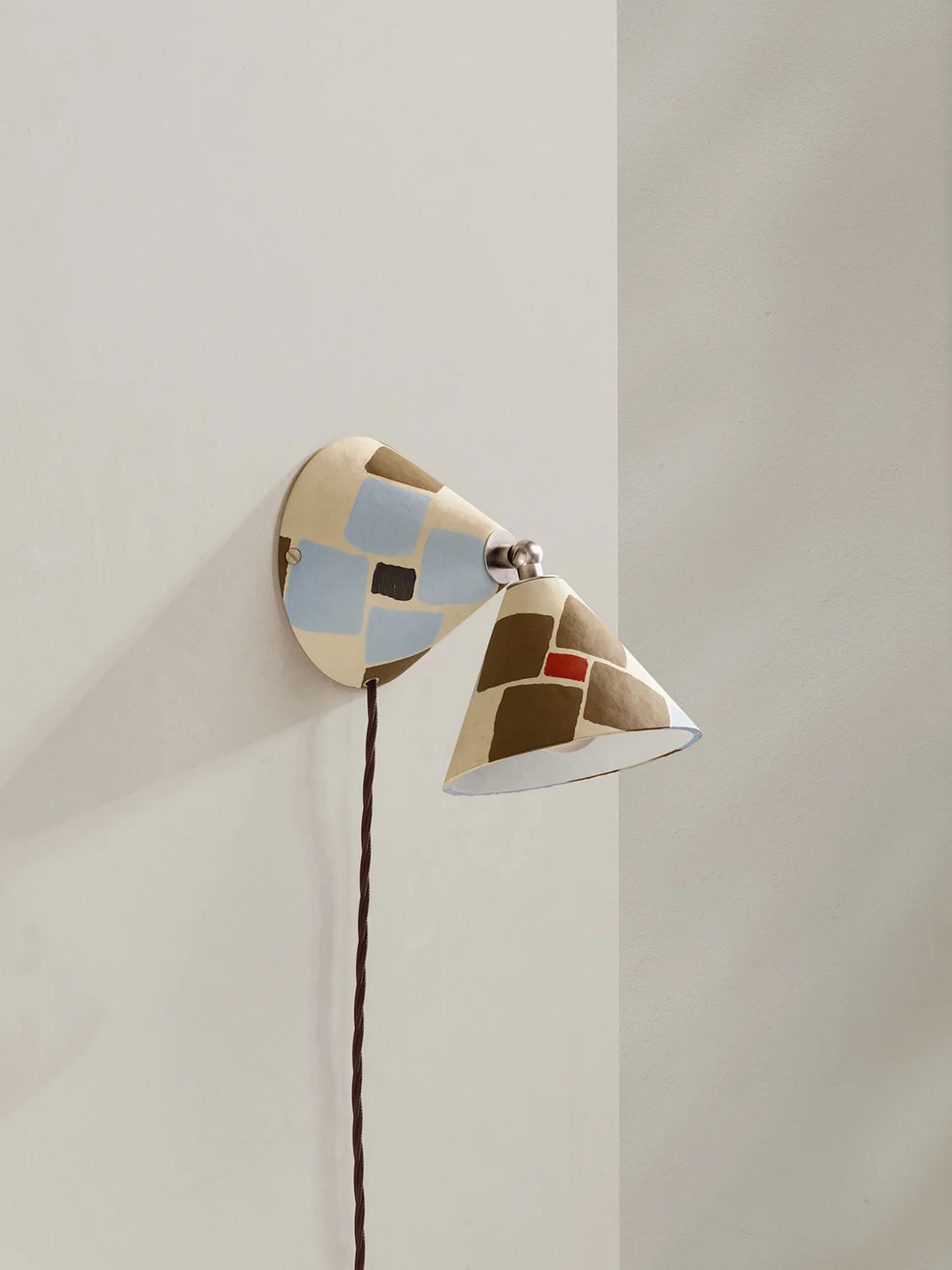 A modern wall-mounted sconce with a dual-cone design, featuring a cream-colored body with geometric patches of light blue, brown, and a small red accent, mounted on a beige wall.