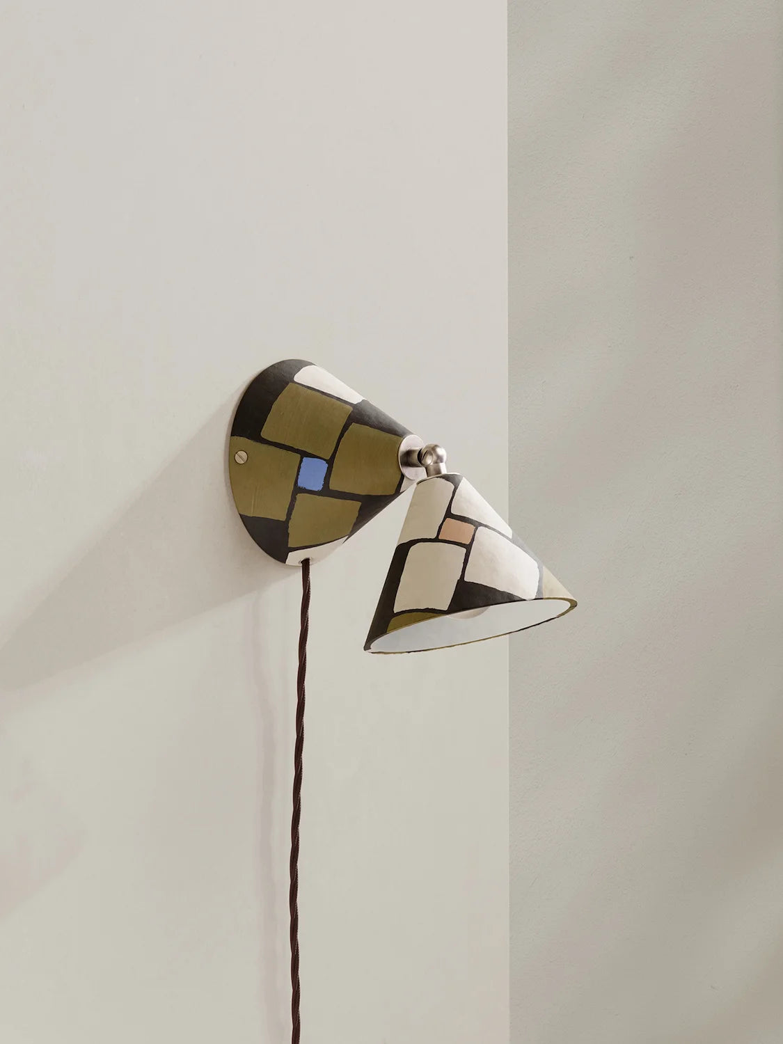 A modern wall-mounted sconce with a geometric mosaic pattern in dark brown, olive green, cream, and a small blue accent, shown mounted on a light beige wall.