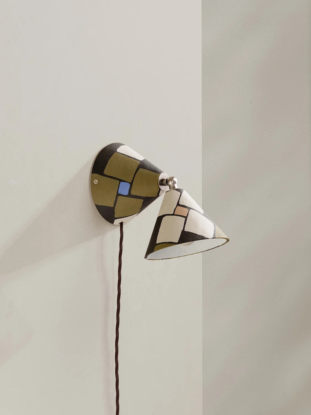 A modern wall-mounted sconce with a geometric mosaic pattern in dark brown, olive green, cream, and a small blue accent, shown mounted on a light beige wall.
