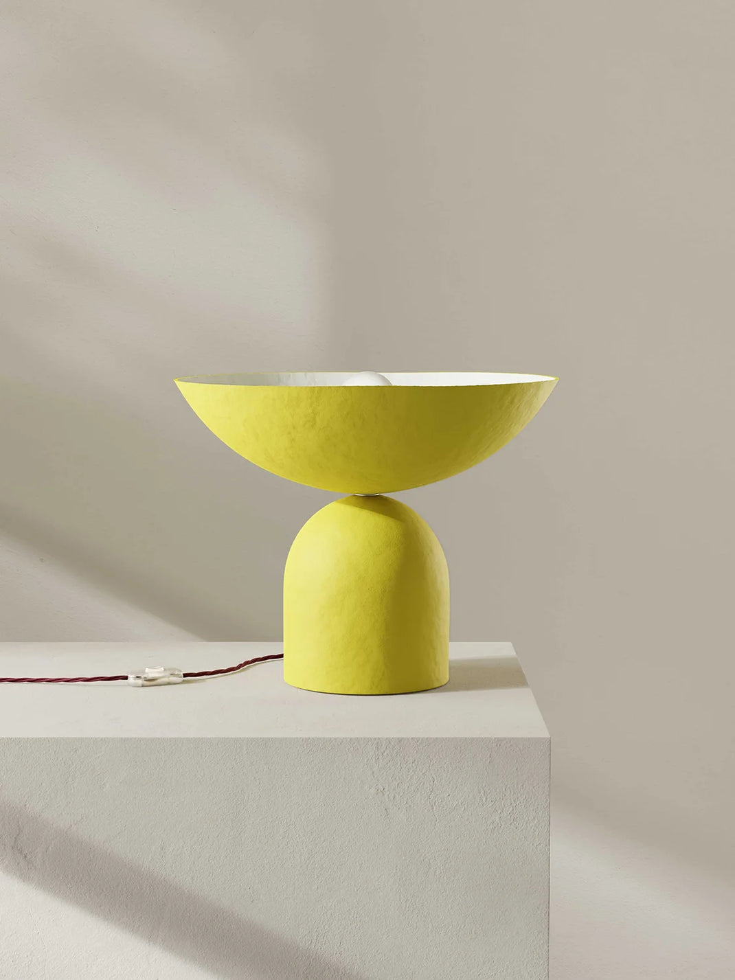 A modern table lamp with a textured bright yellow base and a matching wide, shallow bowl shade, shown on a white pedestal against a neutral beige wall.