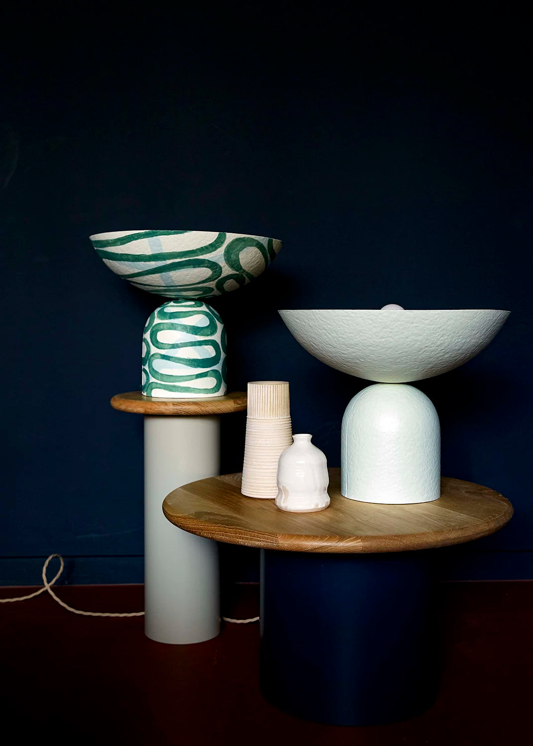 Two uplighter table lamps, one with a green swirl pattern and the other in off-white, on wooden tables against a dark blue wall.