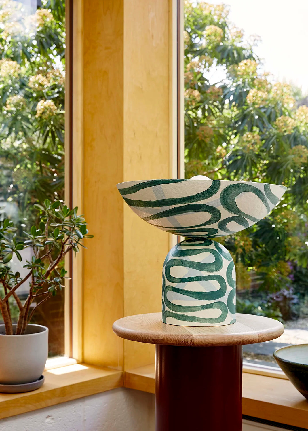 Decorative table lamp with green abstract pattern on a wooden table by a window with greenery outside.