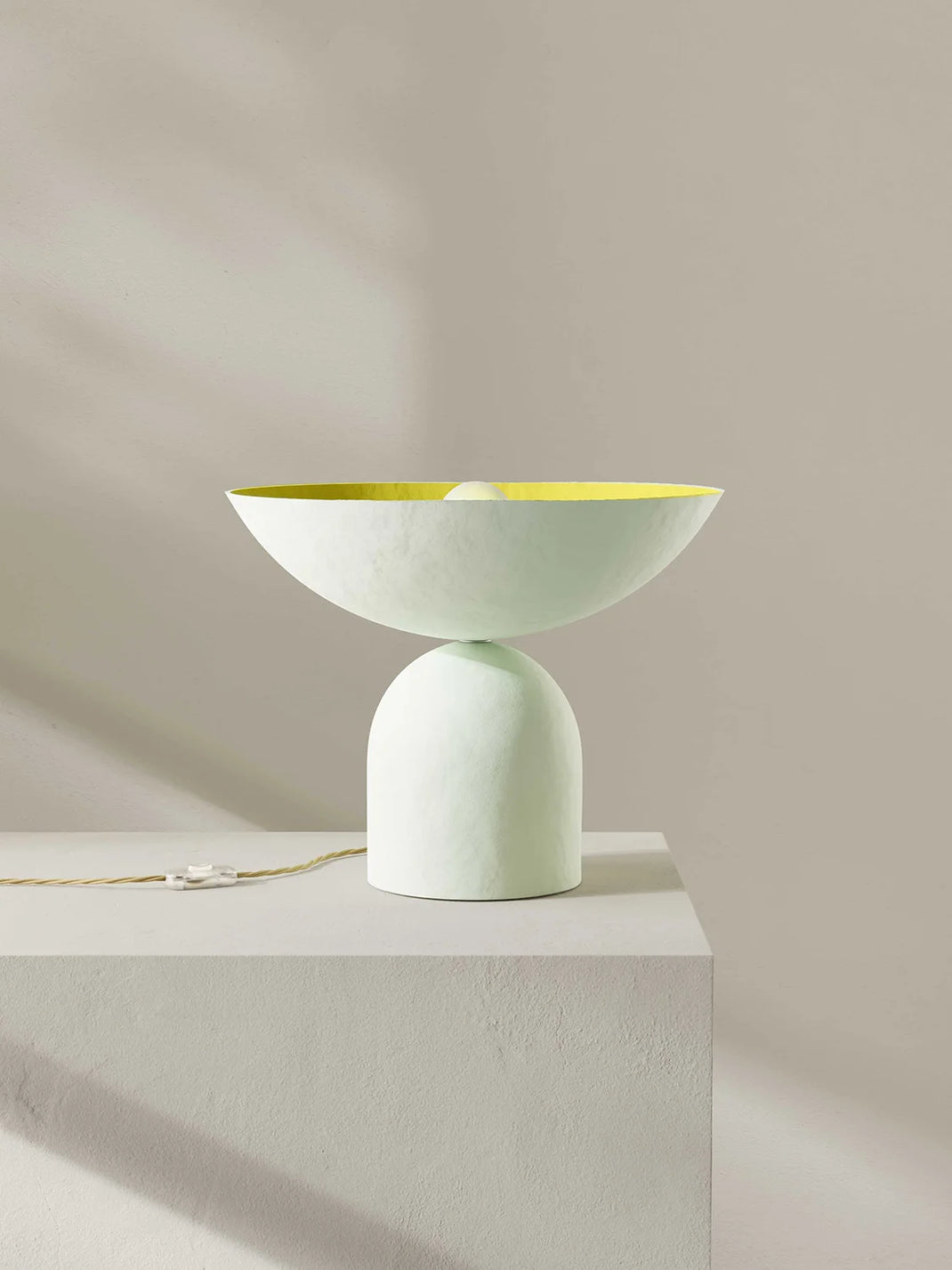 A sculptural table lamp with a wide shallow bowl on a rounded pedestal base, finished in off-white with a bright yellow interior, displayed on a white pedestal against a warm beige wall.