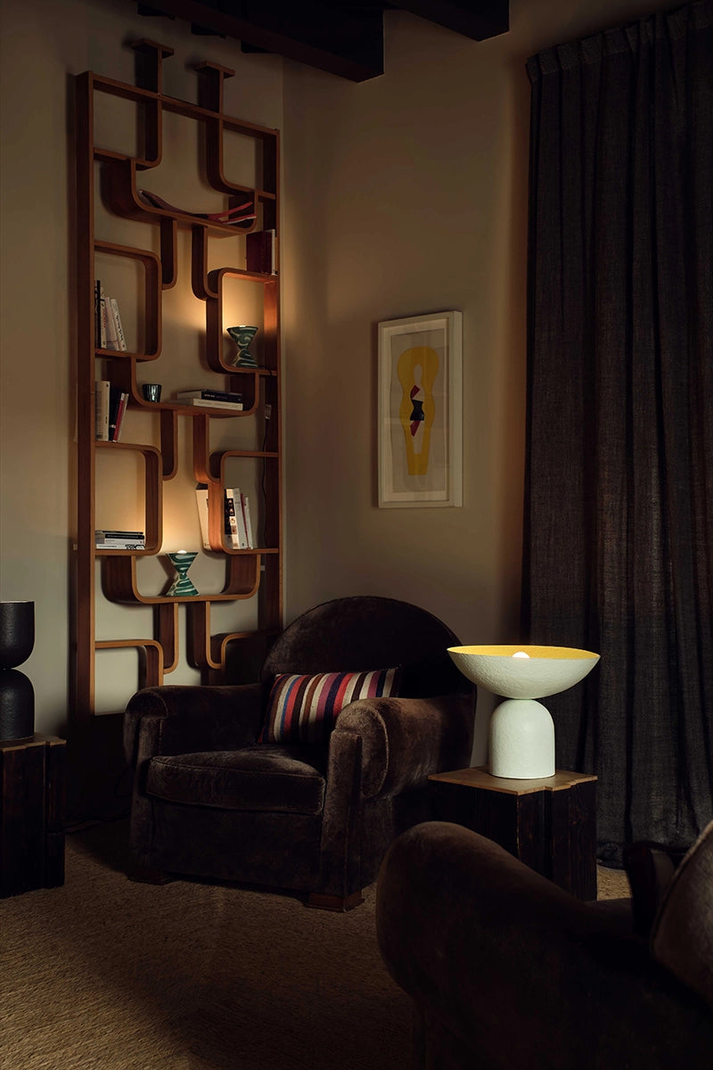 Dark room with an intricate bookshelf, armchair, and decorative items. A white table lamp with yellow interior sits on a side table.