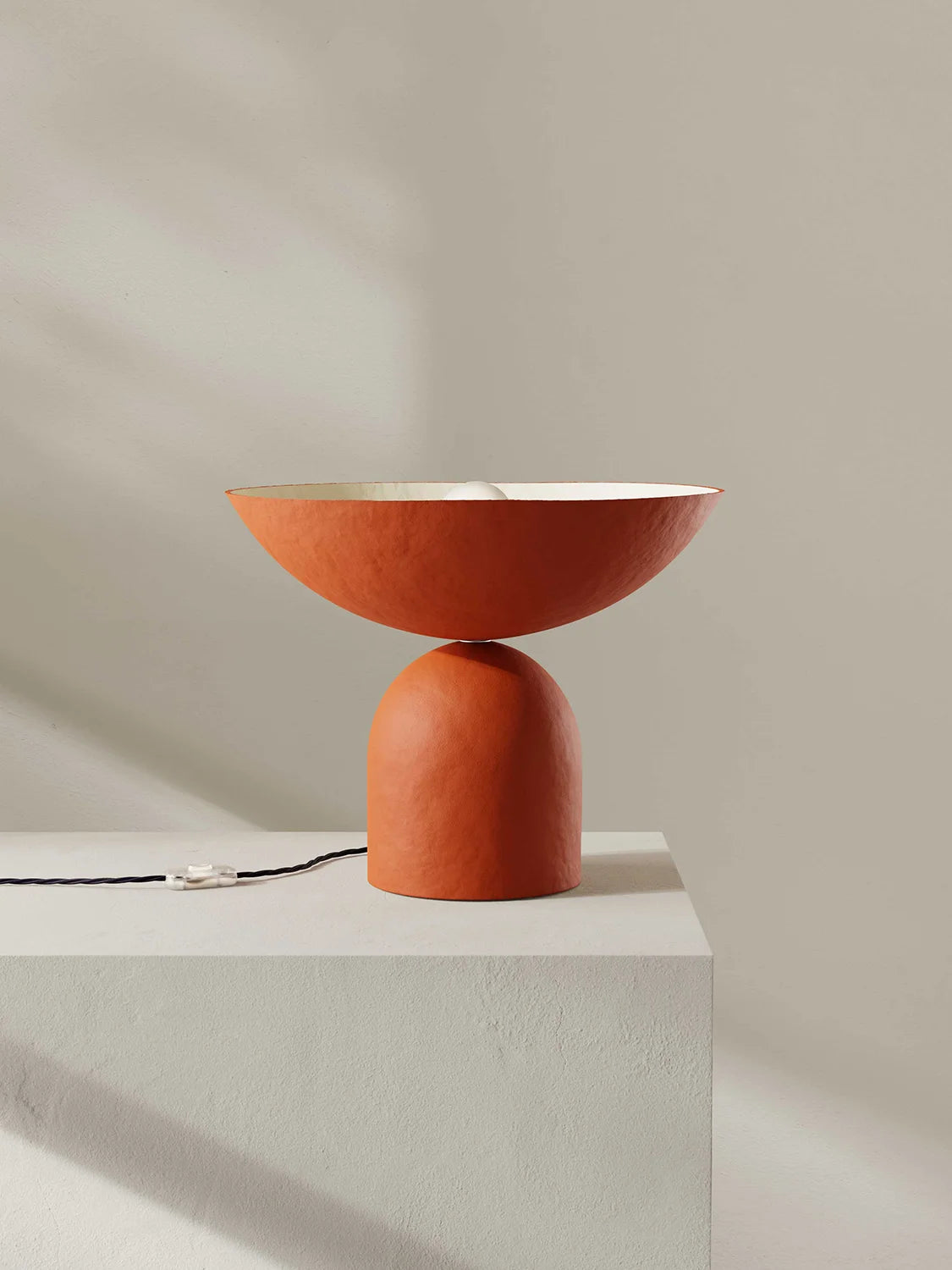 Terracotta-colored table lamp on a light gray pedestal against a beige background