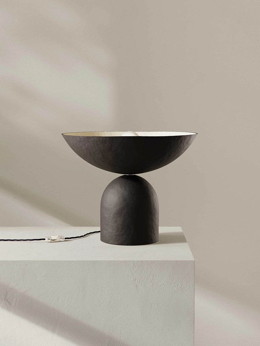 Modern brown-black table lamp with a minimalist design on a light gray pedestal against a beige wall.