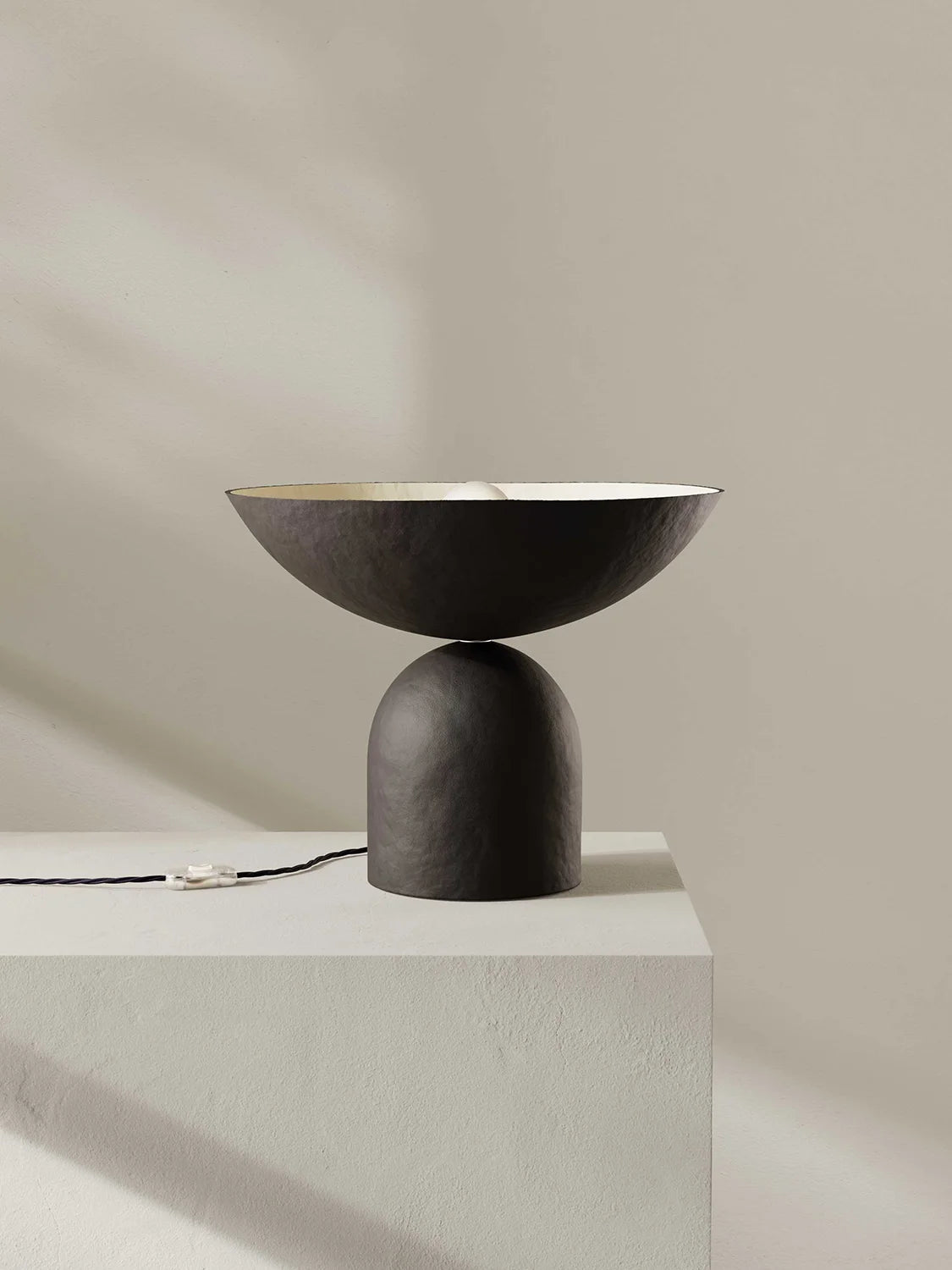 Modern dark brown table lamp with a minimalist design on a light gray pedestal against a beige wall.