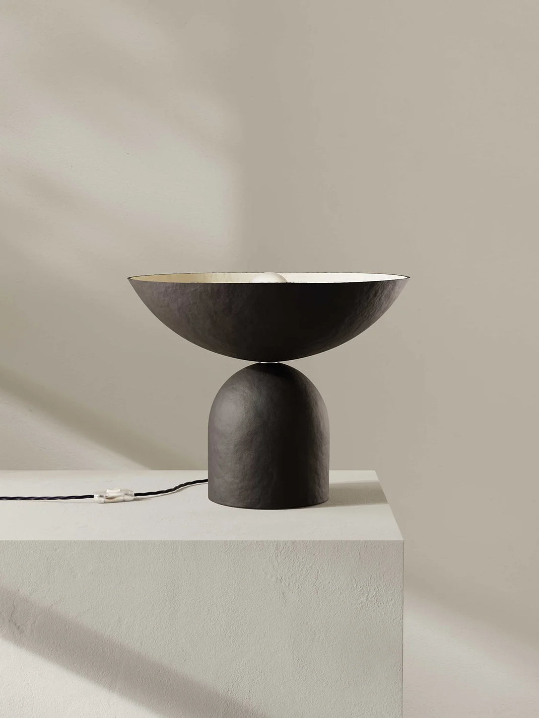 Modern dark brown table lamp with a minimalist design on a light gray pedestal against a beige wall.