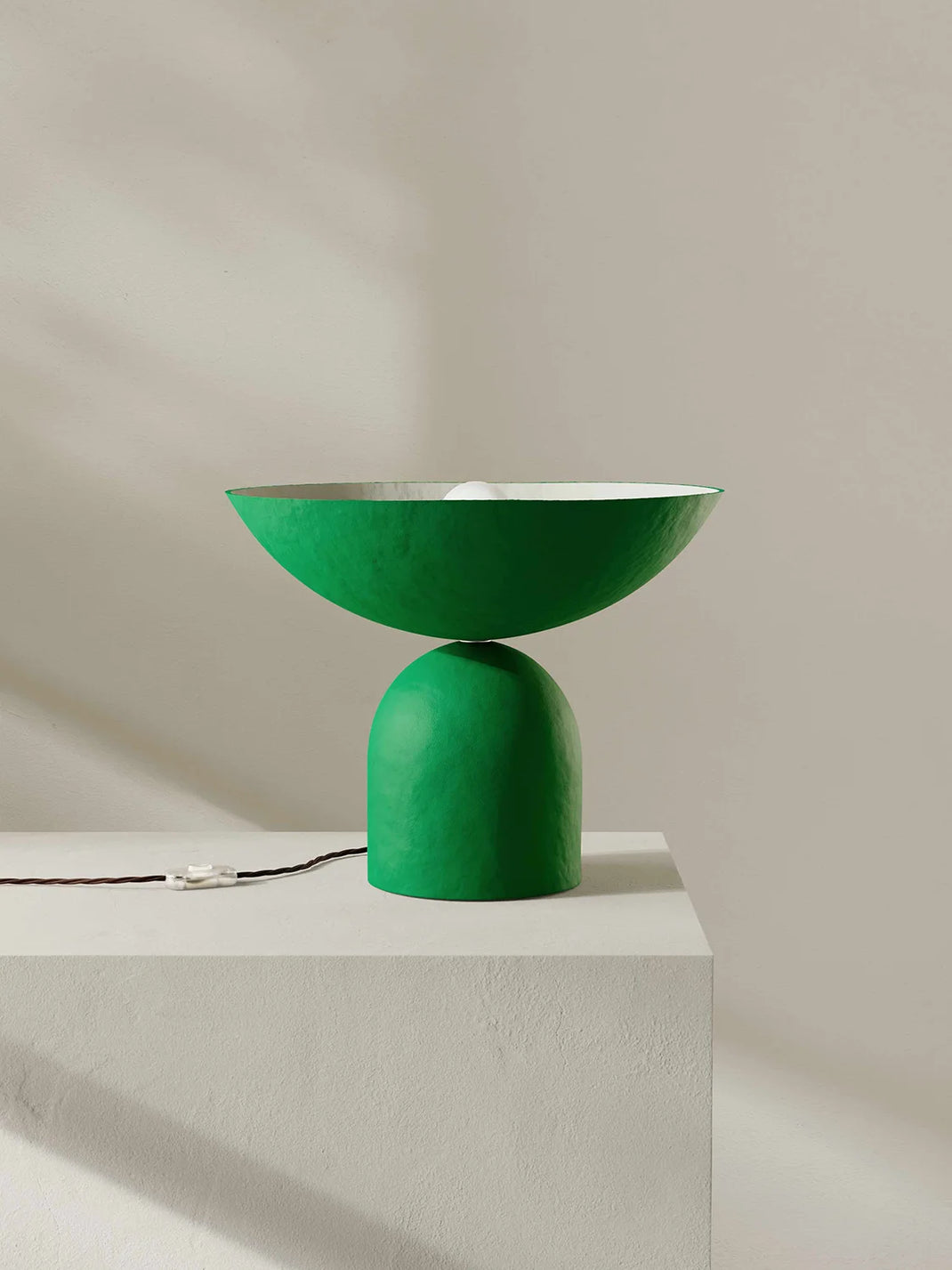 A modern green table lamp with a wide shallow bowl-shaped shade on a rounded cylindrical base, shown on a white pedestal against a warm beige wall with soft shadows.