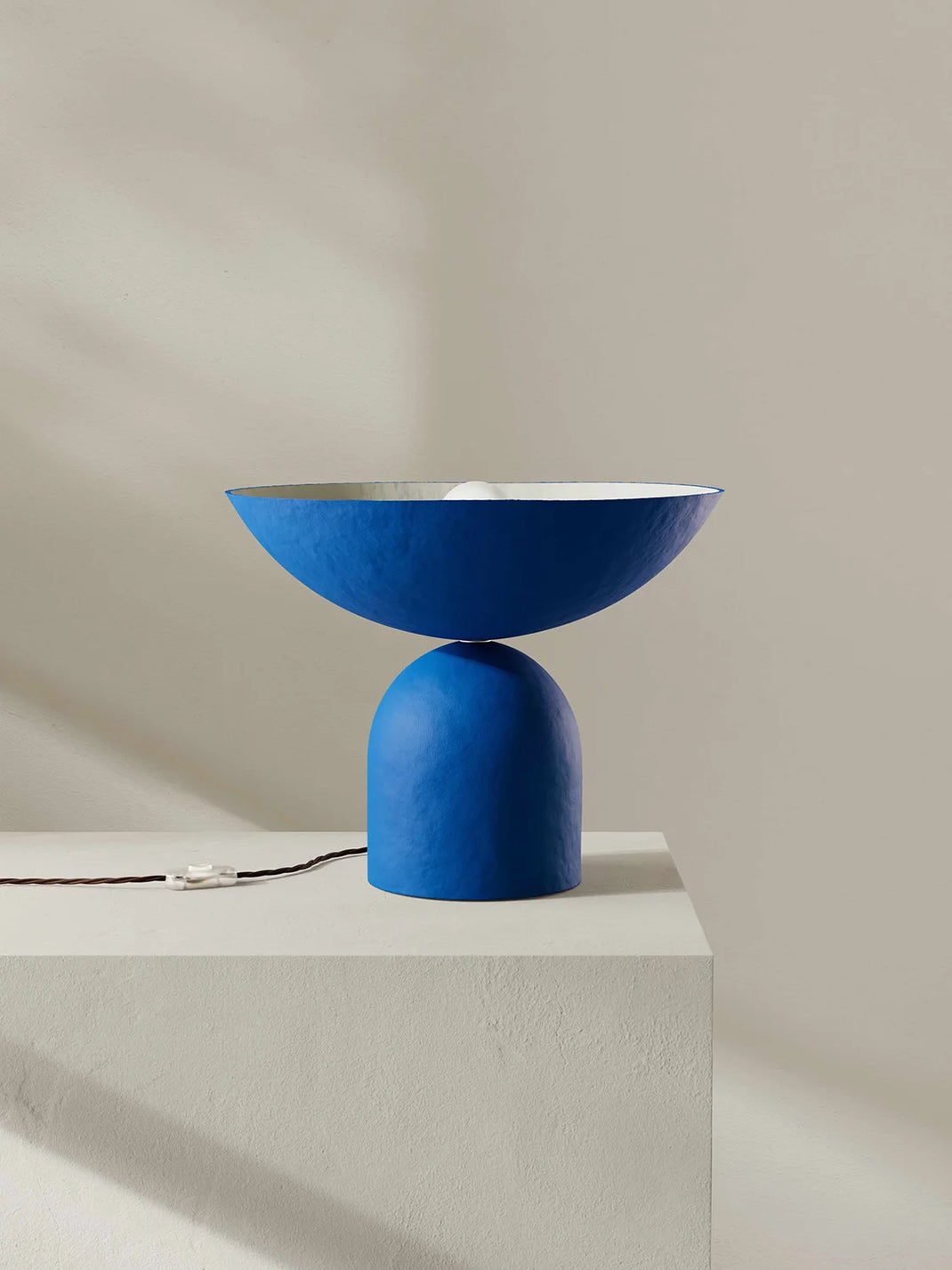 A modern blue table lamp with a wide shallow bowl-shaped shade and a rounded cylindrical base, displayed on a white pedestal against a warm beige wall.