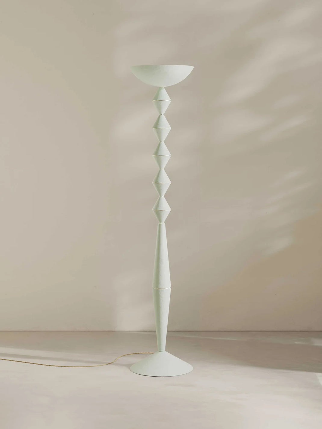 A modern floor lamp with a sculptural, faceted stem and a wide bowl-shaped shade, finished in an off-white color. It stands on a minimalist beige floor against a beige wall with soft, dappled shadows.