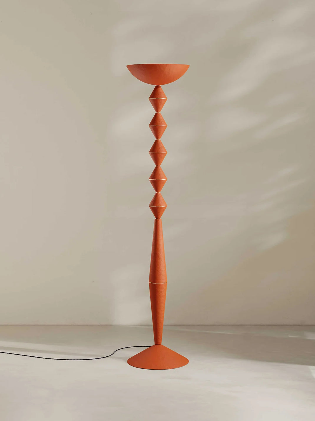 An orange sculptural floor lamp with a bowl-shaped shade and geometric stacked base, standing on a light floor against a beige wall with soft shadow patterns.