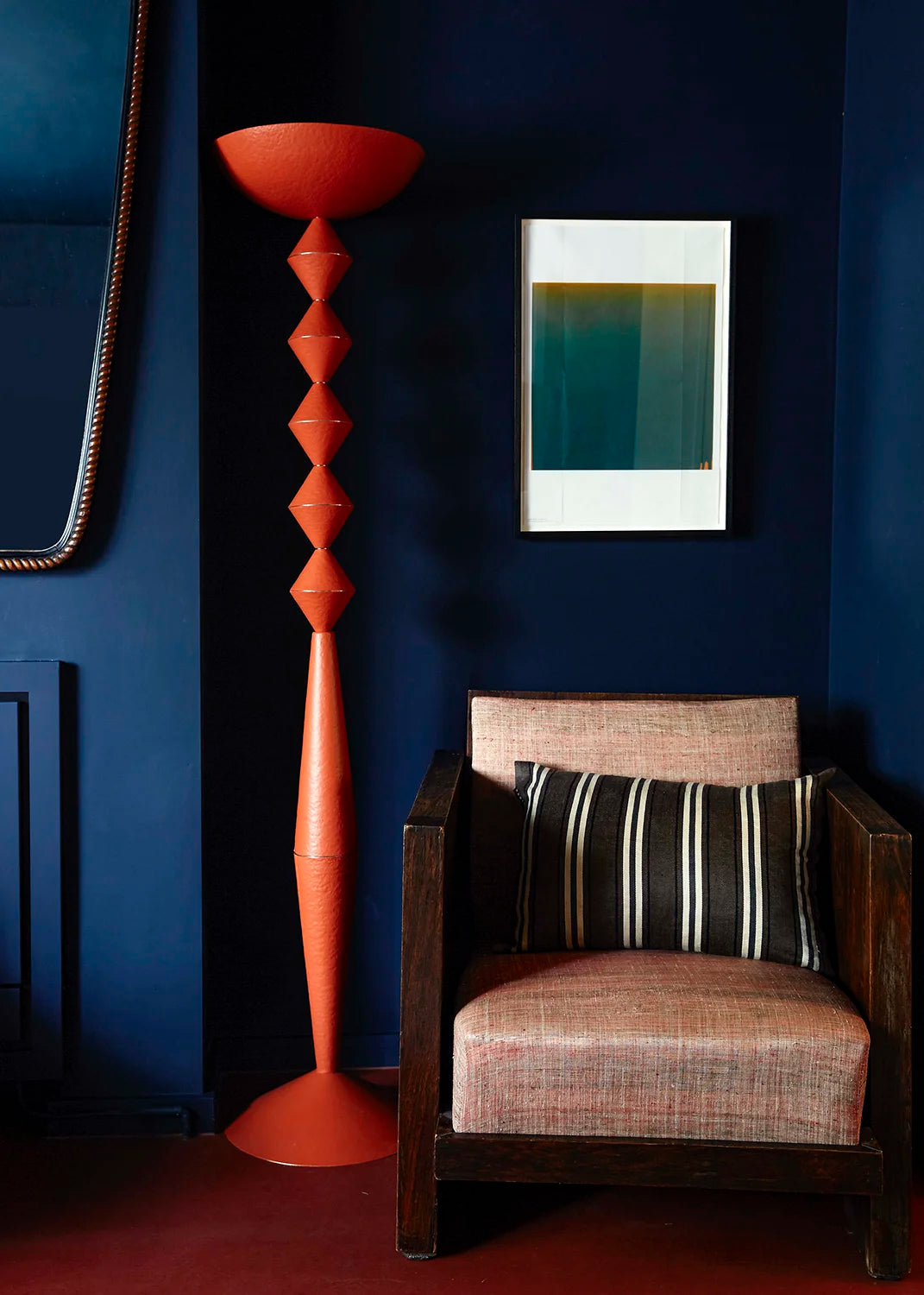 A modern orange sculptural floor lamp stands next to a dark wood armchair with textured blush pink upholstery and a striped pillow, set against a deep navy blue wall with a minimalist art print.