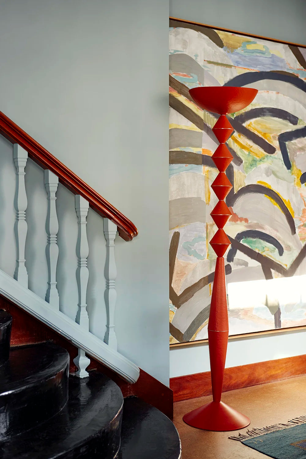 A tall, sculptural red floor lamp with a flared bowl top stands in a modern interior hallway beside a staircase and a large abstract painting.