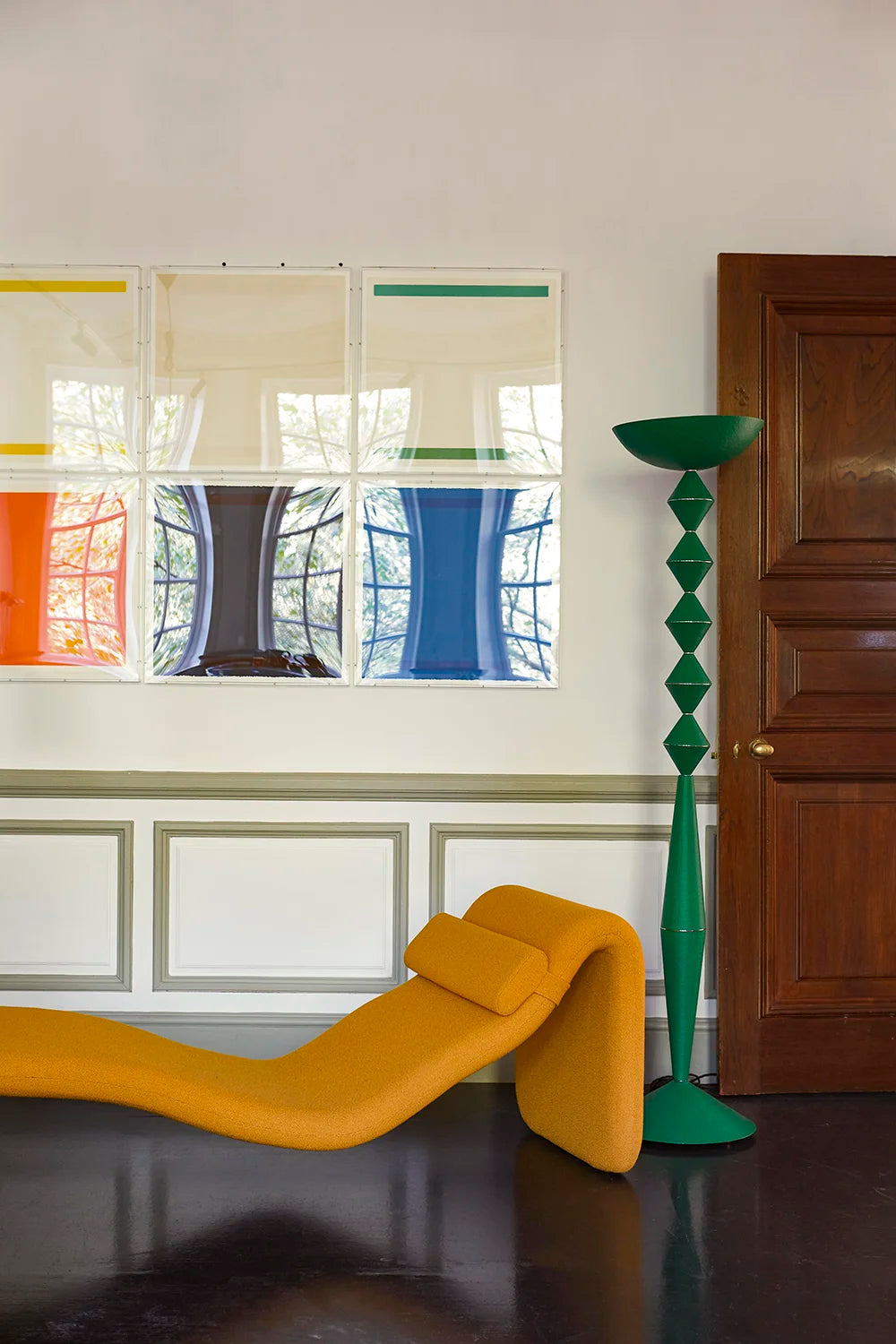A modern interior scene featuring a bright mustard yellow sculptural lounge chair in the foreground, a tall green sculptural floor lamp, and a large abstract multicolor framed artwork on a white paneled wall.