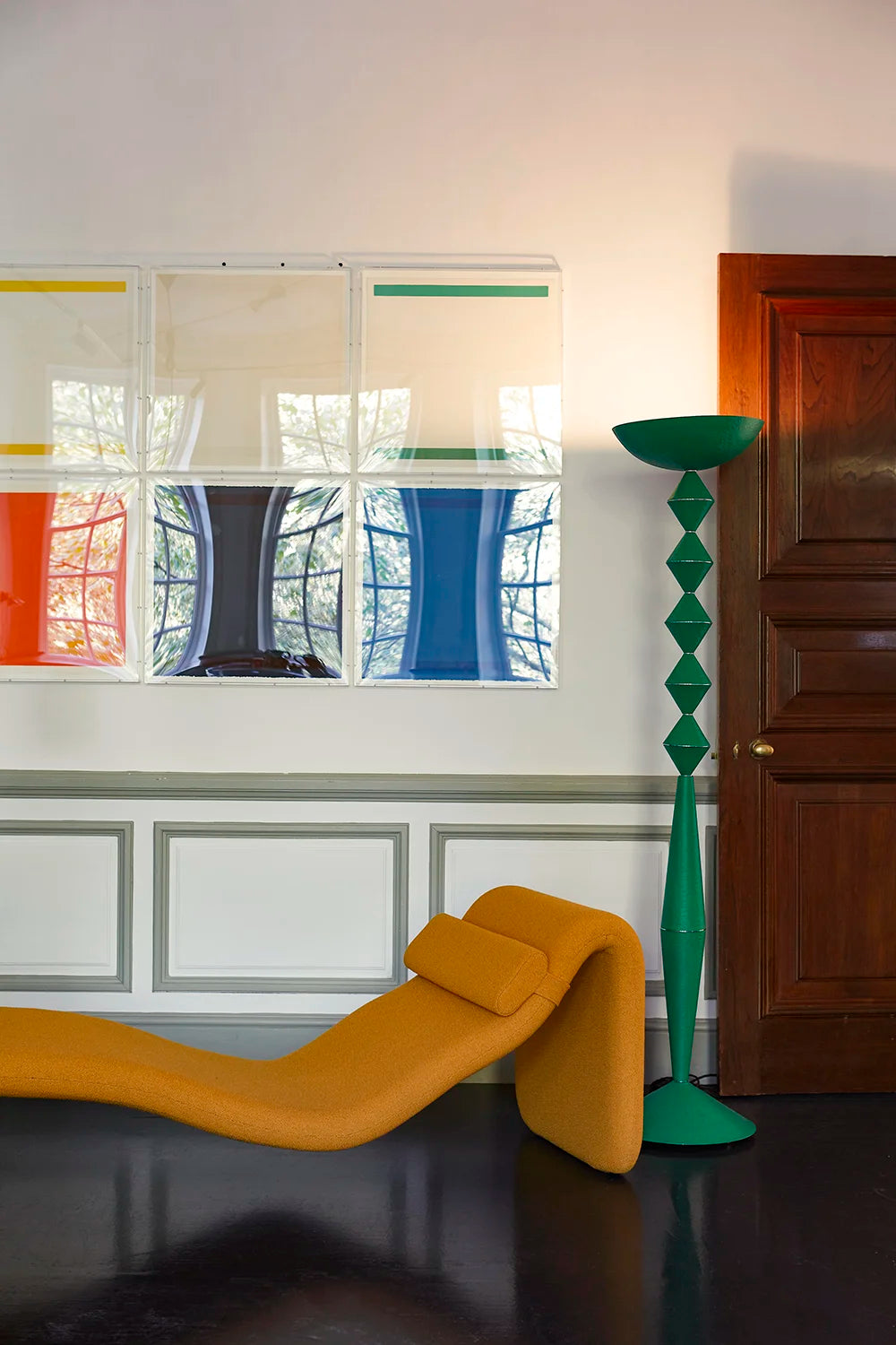 A modern interior scene featuring a bright mustard yellow curved lounge chair in the foreground, a tall green sculptural floor lamp, and a white wall with a colorful abstract framed artwork and a dark wood door.