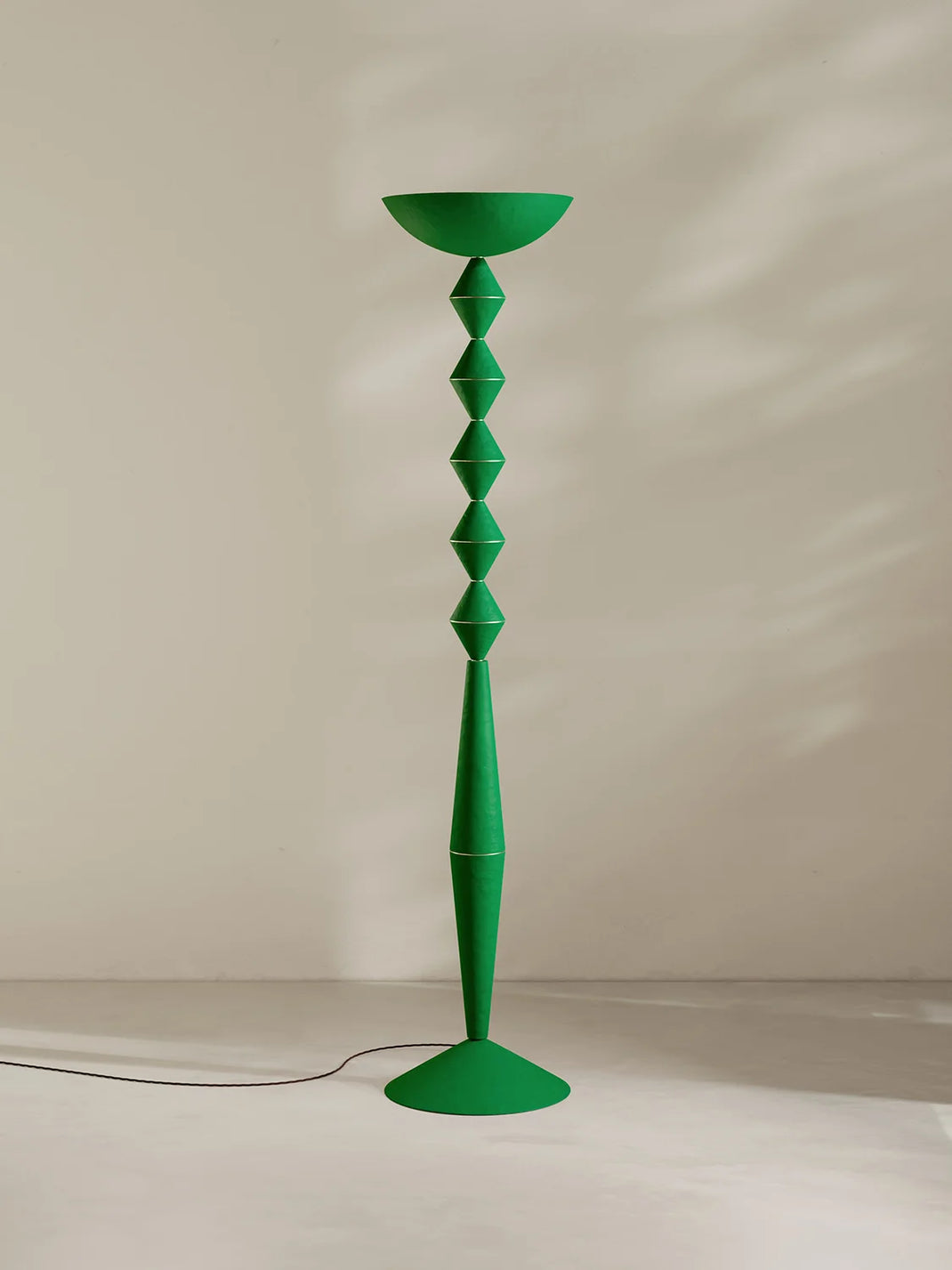 A tall sculptural floor lamp in bright green, featuring a bowl-shaped top, a stacked diamond-patterned stem, and a flared circular base, photographed against a warm off-white wall with soft shadows.