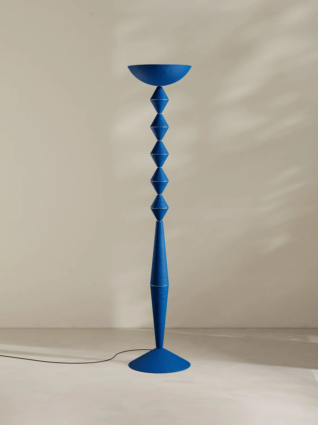 A tall, sculptural floor lamp in a solid medium blue, featuring a wide bowl at the top, a stacked geometric column of diamond-shaped segments, and a flared base. It is photographed in a minimalist studio setting with a warm beige wall and floor, casting a soft shadow.