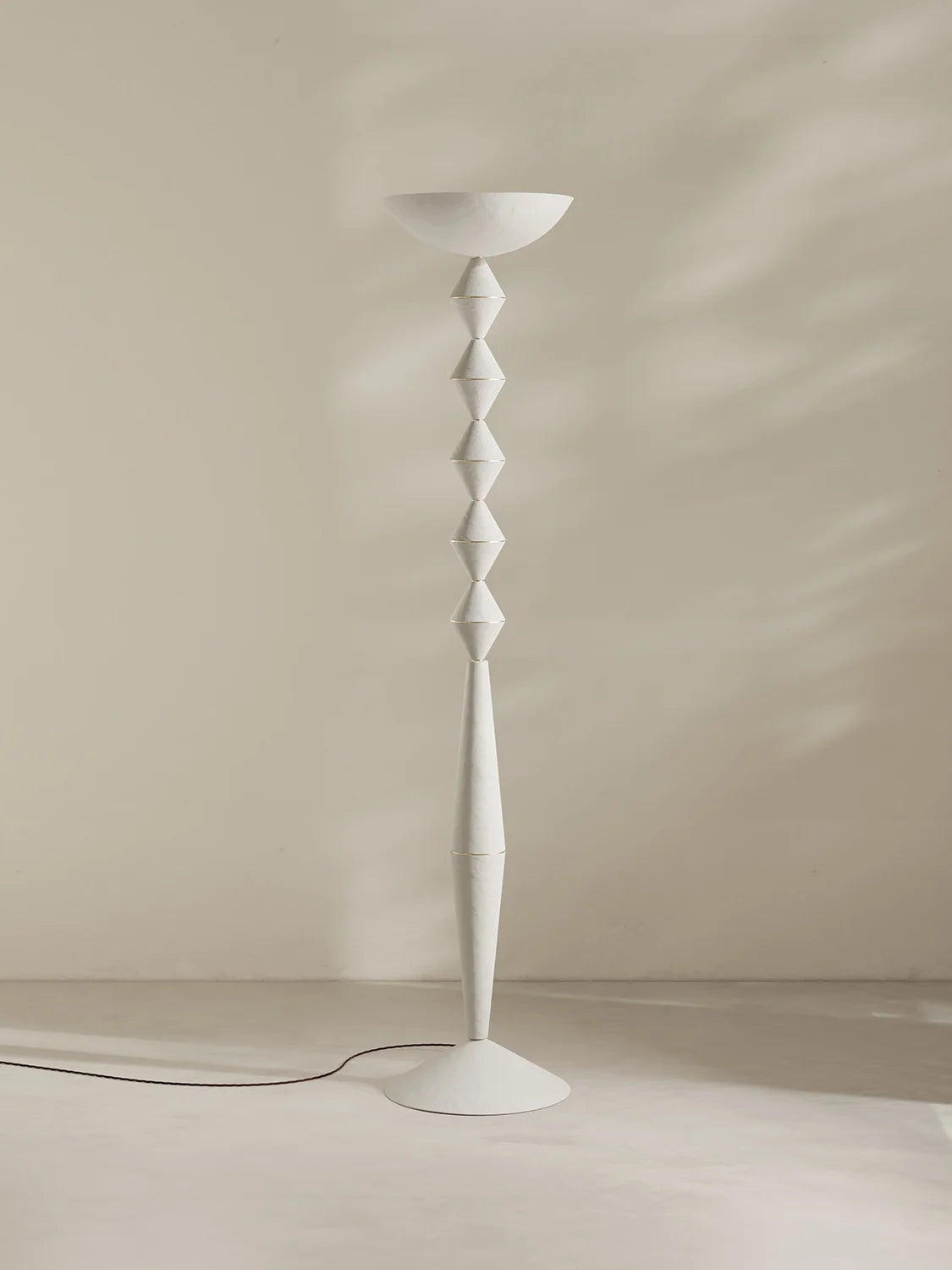 A modern white floor lamp with a sculptural, stacked diamond stem and a shallow bowl-shaped shade, set against a minimalist beige wall with soft shadow patterns.
