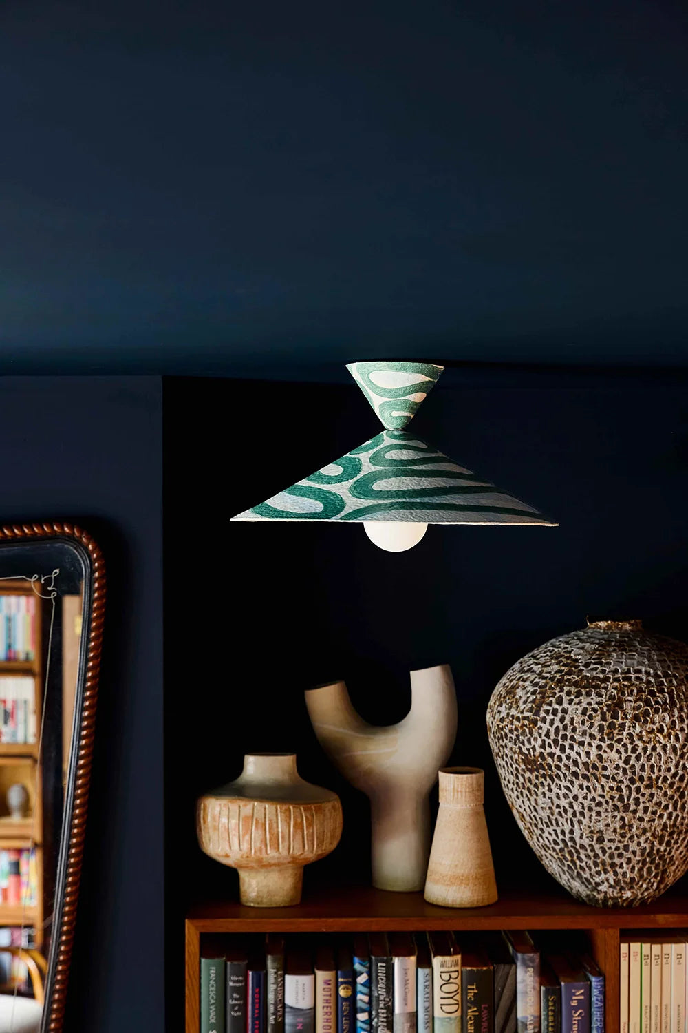 Decorative pendant light with a green and white pattern hanging above a shelf with books and vases.
