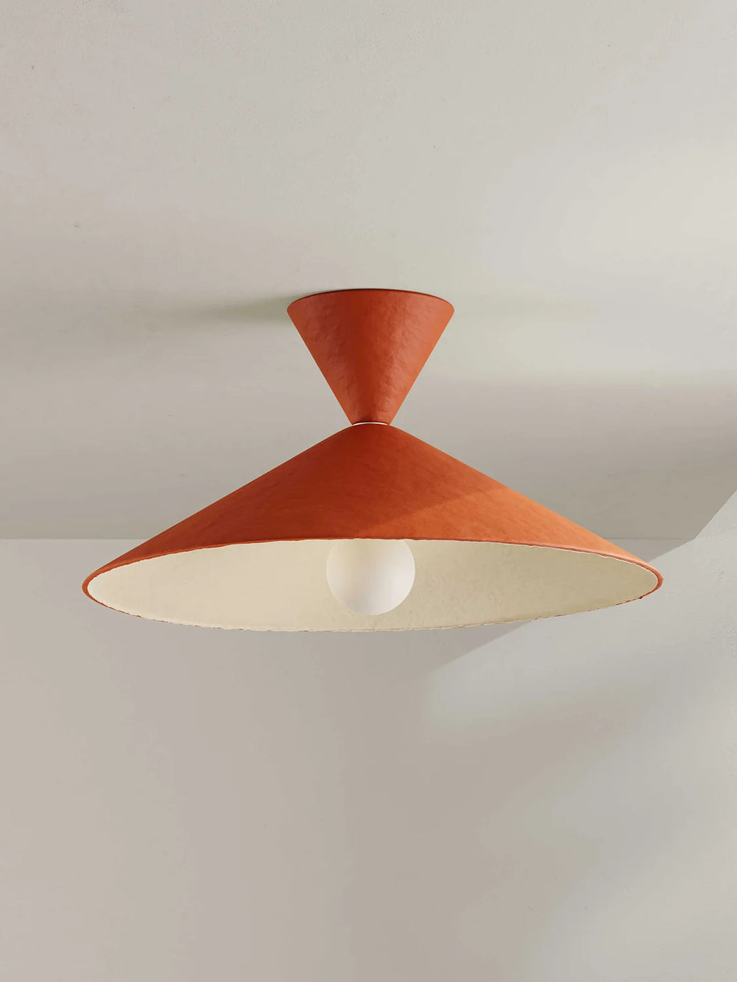 A modern flush mount ceiling light fixture with a terracotta orange conical shade and a white interior, shown against a neutral off-white ceiling.