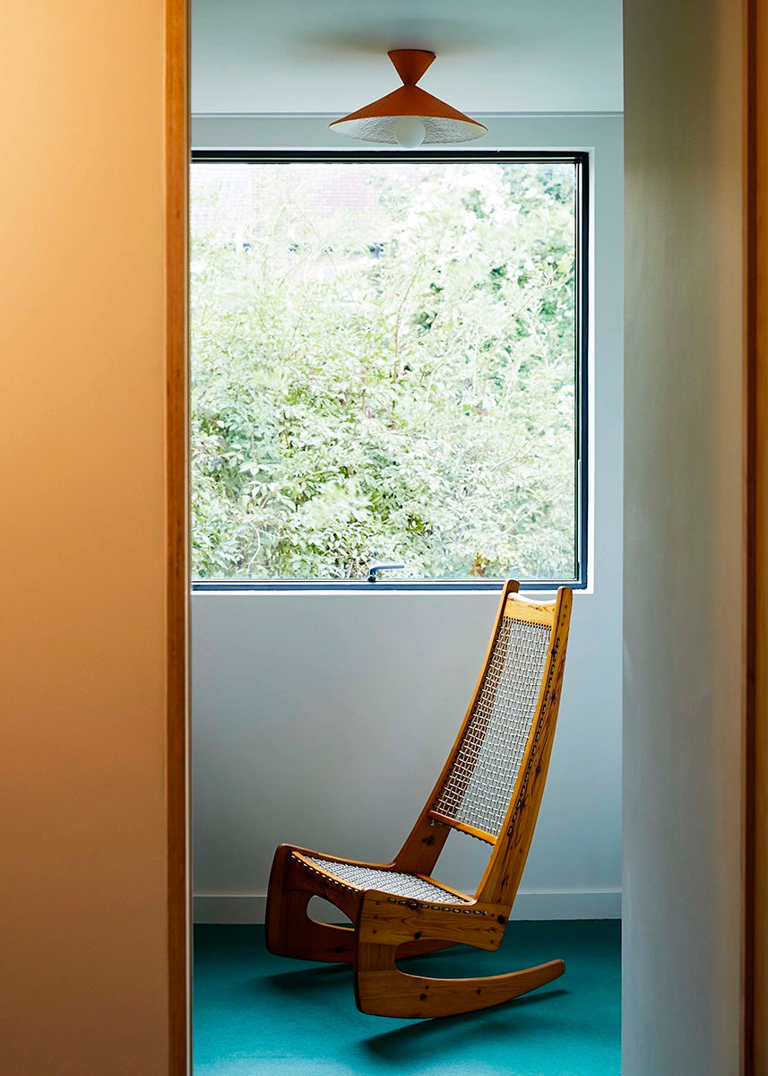 Orange light above a wooden rocking chair in a room with a large window showing greenery outside.