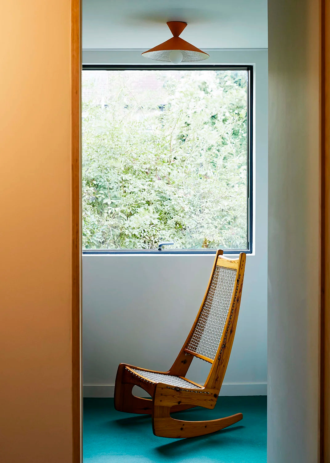 Wooden rocking chair in a room with a large window showing greenery outside. A brick-red ceiling lamp hangs above.