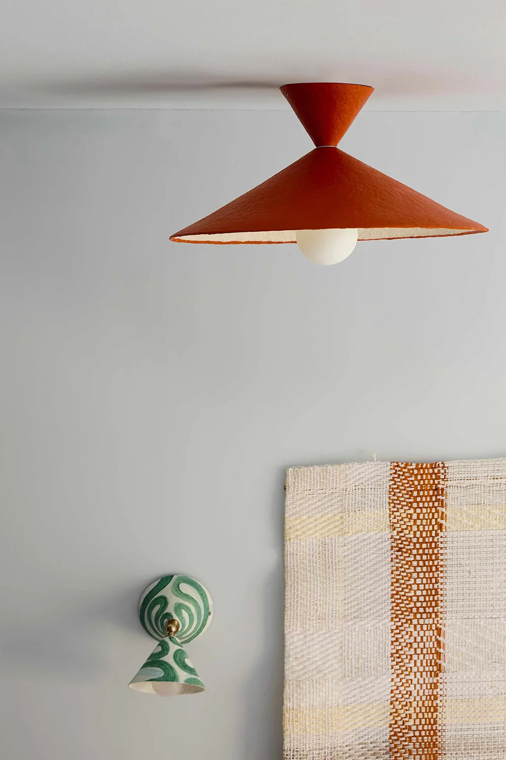 A modern pendant light with a terracotta orange conical shade and a white globe bulb, mounted on a light gray wall above a woven textile. A green and white patterned wall light hangs below the ceiling light.